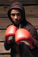 io am pronto per combattere. fiducioso giovane africano uomo nel incappucciato camicia e boxe guanti guardare a telecamera mentre in piedi contro di legno sfondo foto