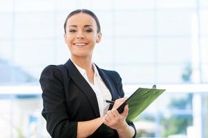 allegro reclutare. bellissimo giovane donna nel vestito formale scrittura qualcosa nel appunti e sorridente mentre in piedi all'aperto foto