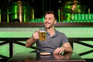 che cosa un' grande giorno allegro giovane uomo Tenere un' boccale con birra e sorridente mentre seduta nel bar foto