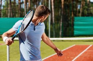 concentrato su gioco. posteriore Visualizza di giovane uomo nel polo camicia giocando tennis mentre in piedi su tennis Tribunale foto