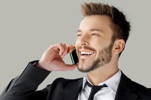 Ciao ritratto di contento giovane uomo nel vestito formale parlando su il Telefono e sorridente mentre in piedi contro grigio sfondo foto