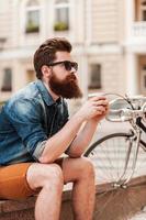 soggiorno calma. riflessivo giovane barbuto uomo Tenere tazza di caffè mentre seduta vicino il suo bicicletta all'aperto foto