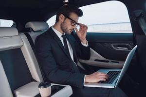 fare surf il rete. bello giovane uomo nel pieno completo da uomo Lavorando utilizzando il computer portatile mentre seduta nel il auto foto