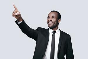 Guarda al di sopra di Là bello giovane africano uomo nel vestito formale puntamento lontano e sorridente mentre in piedi contro grigio sfondo foto