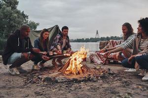 sembra delizioso. gruppo di giovane persone nel casuale indossare torrefazione marshmallows al di sopra di un' fuoco di bivacco mentre riposo vicino il lago foto