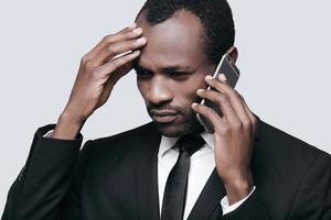 cattivo notizia bello giovane africano uomo toccante testa con mano e parlando su il Telefono mentre in piedi contro grigio sfondo foto
