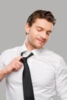 pure tanto pressione. frustrato giovane uomo nel camicia e cravatta toccante slegare il suo cravatta mentre in piedi contro grigio sfondo foto