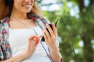 godendo sua preferito musica all'aperto. ritagliata Immagine di bellissimo giovane donna nel cuffie ascoltando per il musica e sorridente mentre pendente a il albero nel un' parco foto