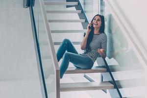 spensierato parlare. bellissimo giovane donna parlando su mobile Telefono e sorridente mentre seduta a il scala a casa foto