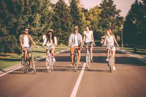 Niente ma amici e strada avanti. gruppo di giovane persone equitazione biciclette lungo un' strada e guardare contento foto