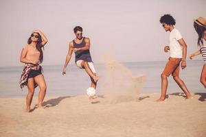 estate divertimento con gli amici. gruppo di allegro giovane persone giocando con calcio palla su il spiaggia con mare nel il sfondo foto