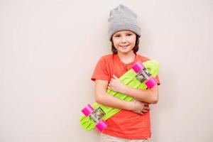 poco pattinatore ragazzo. poco ragazzo nel grigio cappello Tenere colorato skateboard e sorridente mentre in piedi contro grigio sfondo foto