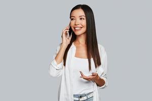 sempre a disposizione. bellissimo giovane asiatico donna parlando su sua inteligente Telefono e sorridente mentre in piedi contro grigio sfondo foto