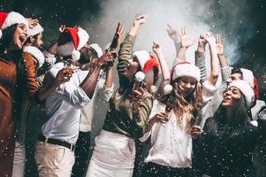 nuovo anno festa. gruppo di bellissimo giovane persone nel Santa cappelli lancio colorato coriandoli e guardare contento foto