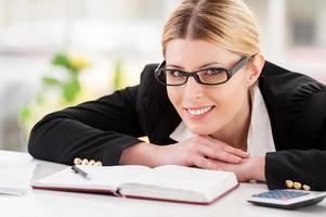 fiducioso attività commerciale signora. bellissimo maturo donna nel vestito formale guardare al di sopra di spalla e sorridente mentre seduta a sua Lavorando posto foto