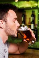 godendo freddo e fresco birra. lato Visualizza di giovane uomo potabile birra mentre seduta a il bar contatore foto