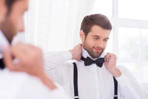 preparazione per un' speciale giorno. bello giovane uomo nel bianca camicia regolazione il suo arco cravatta e sorridente mentre in piedi contro specchio foto