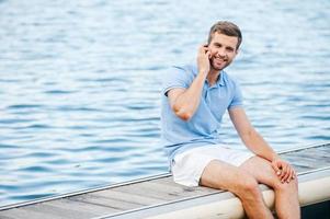 soggiorno collegato ovunque. bello giovane uomo nel polo camicia parlando su il mobile Telefono e sorridente mentre seduta su banchina foto