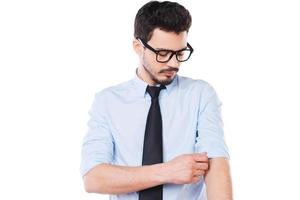 pronto per opera. fiducioso giovane uomo nel camicia e cravatta regolazione il suo manica mentre in piedi contro bianca sfondo foto