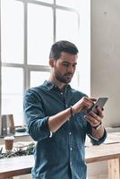 controllo messaggi a partire dal clienti. bello giovane uomo utilizzando inteligente Telefono e sorridente mentre in piedi nel il ufficio foto