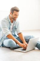 essere a casa nel piacere. bello giovane uomo Lavorando su il computer portatile e sorridente mentre seduta su il pavimento a il suo appartamento foto