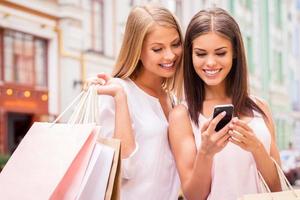 maniaco di shopping gli amici. Due attraente giovane donne Tenere shopping borse e guardare a mobile Telefono insieme mentre in piedi all'aperto foto