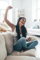 tempo per musica bellissimo giovane donna nel cuffie Tenere inteligente Telefono e Esprimere a gesti mentre seduta su il divano a casa foto