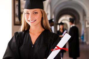 sensazione fiducioso nel sua futuro. contento giovane donna nel la laurea abiti Tenere diploma e sorridente mentre sua amici in piedi nel il sfondo foto