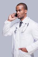 urgente chiamata. fiducioso africano medico parlando su il mobile Telefono e guardare lontano mentre in piedi contro grigio sfondo foto