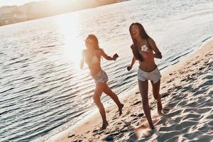contento ragazze. pieno lunghezza di Due attraente giovane donne nel costumi da bagno sorridente mentre in esecuzione su il spiaggia foto