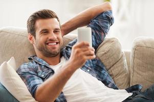 soggiorno nel toccare a casa. allegro giovane uomo Tenere mobile Telefono e sorridente mentre dire bugie su divano foto