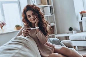 infine incinta. attraente giovane donne guardare a gravidanza test e sorridente mentre seduta su il divano a casa foto