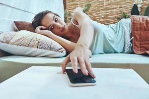 bellissimo giovane donna assunzione sua mobile Telefono mentre dire bugie giù nel domestico letto foto