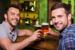 amici potabile birra. Due allegro giovane uomini tostatura con birra e sorridente mentre seduta insieme a il bar contatore foto