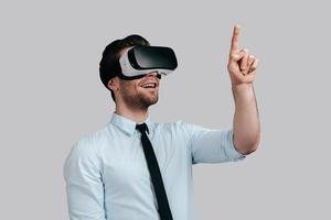 godendo innovativo tecnologie. bene guardare giovane uomo nel virtuale la realtà cuffia puntamento nel il aria mentre in piedi contro grigio sfondo foto
