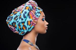 fiducia e bellezza. lato Visualizza di bellissimo africano donna indossare un' foulard e conservazione occhi chiuso mentre in piedi contro nero sfondo foto