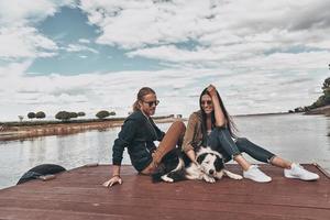 con loro migliore amico. bellissimo giovane coppia sorridente mentre seduta su il di legno piattaforma vicino il fiume foto