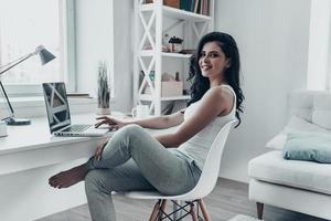 fare surf il netto a casa. bellissimo giovane donna utilizzando il computer portatile e guardare a telecamera con Sorridi mentre seduta a il scrivania a casa foto