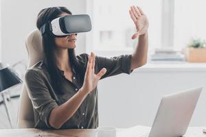 donna con occhiali vr. giovane donna sicura di sé in cuffia per realtà virtuale che punta in aria mentre è seduta al suo posto di lavoro in ufficio
