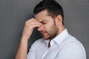 sensazione stanco e depresso. frustrato giovane uomo toccante il suo viso con mano e conservazione occhi chiuso mentre in piedi contro grigio sfondo foto