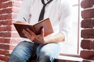 scrittura giù il suo pensieri. avvicinamento di giovane uomo nel camicia e cravatta scrittura qualcosa nel Nota tampone mentre seduta a il finestra davanzale foto