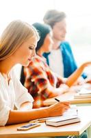 Lavorando difficile per un' bene futuro. concentrato giovane donna scrittura nel Nota tampone mentre seduta a sua scrivania nel il aula foto