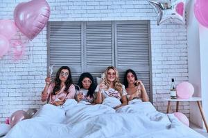 così tanto divertimento quattro attraente giovane donne nel pigiama potabile cocktail mentre dire bugie nel il letto con palloncini tutti al di sopra di il camera foto
