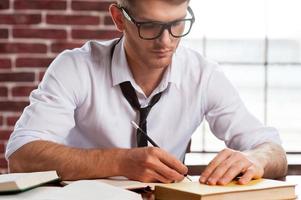 fiducioso e creativo. bello giovane uomo nel camicia e cravatta scrittura qualcosa nel Nota tampone mentre seduta a il suo Lavorando posto foto