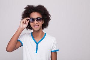 soggiorno freddo allegro adolescenziale africano ragazzo regolazione il suo occhiali da sole e sorridente mentre in piedi isolato su grigio sfondo foto