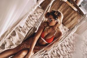 godendo caldo giorno. superiore Visualizza di attraente giovane donna nel rosso bikini riposo mentre seduta nel amaca su il spiaggia foto