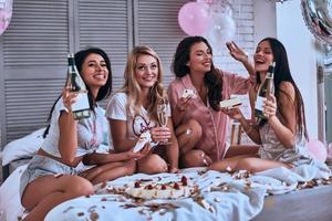 indimenticabile festa. quattro bellissimo giovane donne nel pigiama mangiare torta e potabile Champagne mentre avendo un' sonno festa foto
