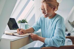 bellissimo giovane sorridente donna scrittura qualcosa nel Nota tampone mentre Lavorando a casa foto