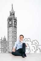 sognare di Londra. giovane bello uomo conservazione mano su mento e guardare lontano con Sorridi mentre seduta su il pavimento con illustrazione di grande Ben a il sfondo foto