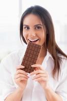 ragazza godendo cioccolato. allegro giovane sorridente donna nel bianca camicia mangiare cioccolato bar e guardare a telecamera foto